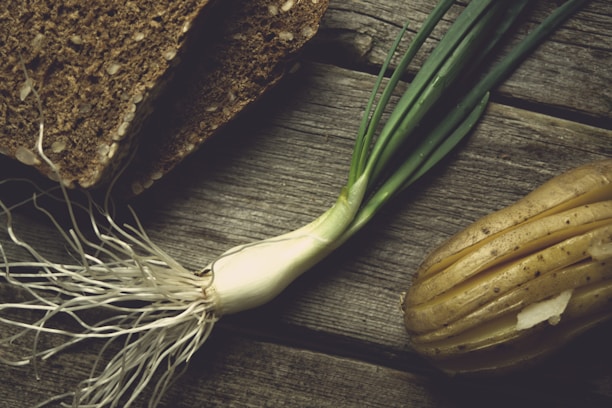 A rustic wooden surface supports slices of whole-grain bread, a bunch of fresh green onions with roots, and a sliced, seasoned potato. The composition suggests a natural, earthy atmosphere.