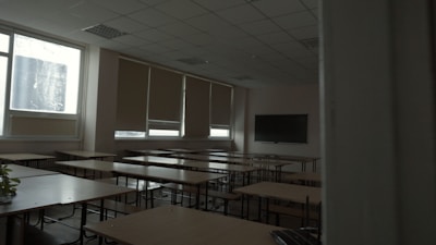 A dimly lit classroom with several rows of empty wooden desks and chairs. The windows on the side have blinds partially drawn, letting in minimal natural light. A large blackboard is mounted on the beige walls at the front of the room.