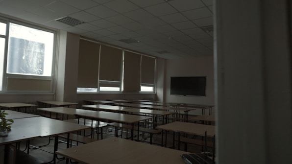 A dimly lit classroom with several rows of empty wooden desks and chairs. The windows on the side have blinds partially drawn, letting in minimal natural light. A large blackboard is mounted on the beige walls at the front of the room.