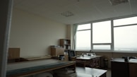 A spacious, well-lit office with large windows providing a view of the outdoors. The room features multiple desks with chairs, bookshelves, and stacks of papers. The ceiling is lined with panels, and the walls are neutral in color. The overall setup gives a neat and organized appearance.