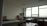 A spacious, well-lit office with large windows providing a view of the outdoors. The room features multiple desks with chairs, bookshelves, and stacks of papers. The ceiling is lined with panels, and the walls are neutral in color. The overall setup gives a neat and organized appearance.