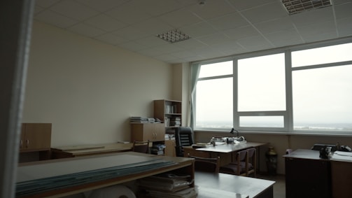 A spacious, well-lit office with large windows providing a view of the outdoors. The room features multiple desks with chairs, bookshelves, and stacks of papers. The ceiling is lined with panels, and the walls are neutral in color. The overall setup gives a neat and organized appearance.