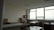 A spacious, well-lit office with large windows providing a view of the outdoors. The room features multiple desks with chairs, bookshelves, and stacks of papers. The ceiling is lined with panels, and the walls are neutral in color. The overall setup gives a neat and organized appearance.