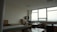 A spacious, well-lit office with large windows providing a view of the outdoors. The room features multiple desks with chairs, bookshelves, and stacks of papers. The ceiling is lined with panels, and the walls are neutral in color. The overall setup gives a neat and organized appearance.