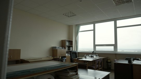 A spacious, well-lit office with large windows providing a view of the outdoors. The room features multiple desks with chairs, bookshelves, and stacks of papers. The ceiling is lined with panels, and the walls are neutral in color. The overall setup gives a neat and organized appearance.