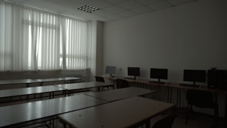 A dimly lit classroom or computer lab with several empty desks and a row of computer monitors along one wall. The window has closed vertical blinds, allowing minimal light to filter through.