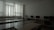 A dimly lit classroom or computer lab with several empty desks and a row of computer monitors along one wall. The window has closed vertical blinds, allowing minimal light to filter through.