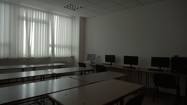A dimly lit classroom or computer lab with several empty desks and a row of computer monitors along one wall. The window has closed vertical blinds, allowing minimal light to filter through.