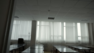 A large room with multiple tables aligned in rows and large windows covered with vertical blinds. The room is empty, with stacked chairs and a stand visible in the corner. The lighting is predominantly soft and natural, coming through the windows.