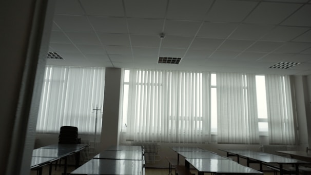 A large room with multiple tables aligned in rows and large windows covered with vertical blinds. The room is empty, with stacked chairs and a stand visible in the corner. The lighting is predominantly soft and natural, coming through the windows.