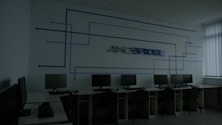 A dimly lit computer lab features several workstations with monitors and keyboards arranged in a line. A design composed of blue lines and the word 'Amcbridge' is visible on the wall. Vertical blinds partially cover the window, letting in minimal light.