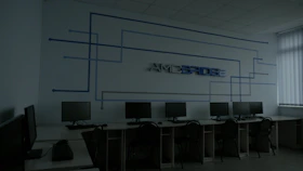 A dimly lit computer lab features several workstations with monitors and keyboards arranged in a line. A design composed of blue lines and the word 'Amcbridge' is visible on the wall. Vertical blinds partially cover the window, letting in minimal light.