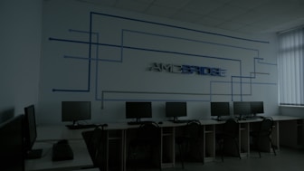 A dimly lit computer lab features several workstations with monitors and keyboards arranged in a line. A design composed of blue lines and the word 'Amcbridge' is visible on the wall. Vertical blinds partially cover the window, letting in minimal light.