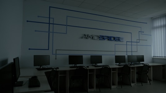 A dimly lit computer lab features several workstations with monitors and keyboards arranged in a line. A design composed of blue lines and the word 'Amcbridge' is visible on the wall. Vertical blinds partially cover the window, letting in minimal light.