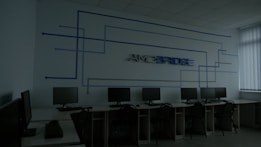 A dimly lit computer lab features several workstations with monitors and keyboards arranged in a line. A design composed of blue lines and the word 'Amcbridge' is visible on the wall. Vertical blinds partially cover the window, letting in minimal light.