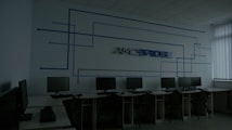 A dimly lit computer lab features several workstations with monitors and keyboards arranged in a line. A design composed of blue lines and the word 'Amcbridge' is visible on the wall. Vertical blinds partially cover the window, letting in minimal light.