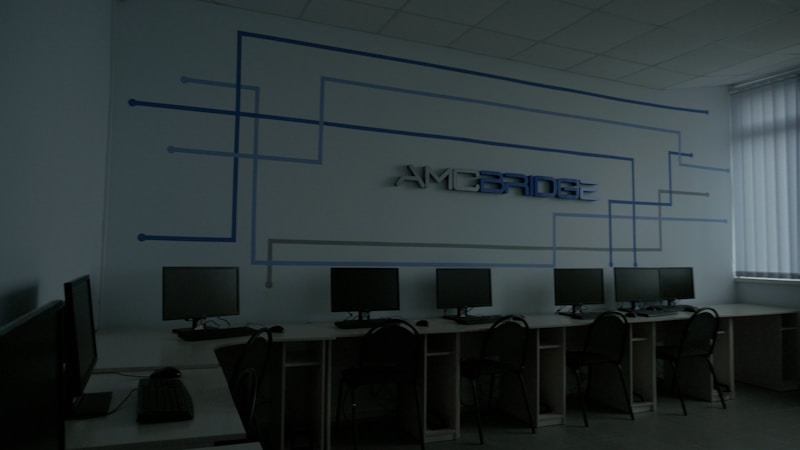 A dimly lit computer lab features several workstations with monitors and keyboards arranged in a line. A design composed of blue lines and the word 'Amcbridge' is visible on the wall. Vertical blinds partially cover the window, letting in minimal light.