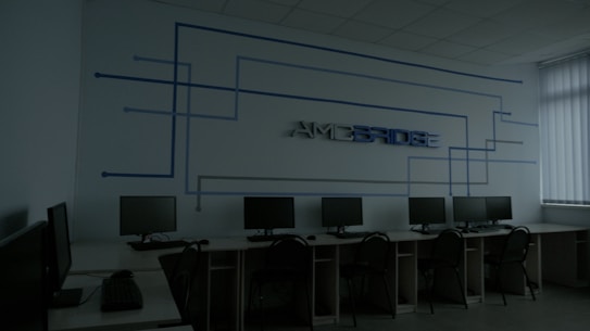 A dimly lit computer lab features several workstations with monitors and keyboards arranged in a line. A design composed of blue lines and the word 'Amcbridge' is visible on the wall. Vertical blinds partially cover the window, letting in minimal light.