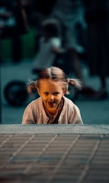 A young child with braided hair leans forward playfully, sticking out their tongue. The background is blurred, suggesting a bustling outdoor area with other people and possibly a bicycle.