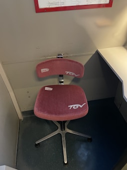 A chair with a red cushioned seat and backrest, labeled with 'TGV' and 'SNCF' logos, is positioned in a small, enclosed space near a desk. The chair has a metal base with a swivel feature. Above the chair, a framed schedule or chart is mounted on the wall.