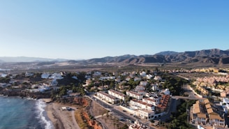 A coastal town featuring a blend of residential buildings with white and red-tiled roofs, nestled against a mountainous backdrop. The shoreline is marked by a sandy beach meeting the blue sea, and the terrain transitions into arid, brown hills. Streets and a few scattered green patches of trees are visible.