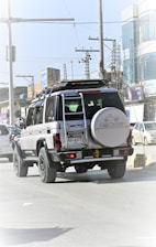A black and white Toyota Land Cruiser is driving on a city street. It has a spare tire mounted on the back and is surrounded by other vehicles. The street is lined with buildings, power lines, and various urban elements in the background.
