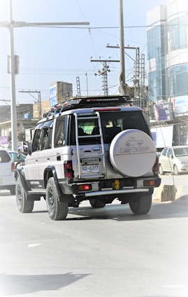 A black and white Toyota Land Cruiser is driving on a city street. It has a spare tire mounted on the back and is surrounded by other vehicles. The street is lined with buildings, power lines, and various urban elements in the background.