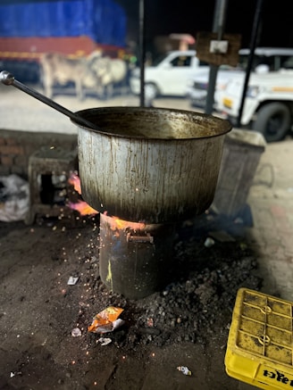 A large, metal pot is placed on a stove or makeshift burner with wood burning underneath. The surrounding area is dimly lit, suggesting an outdoor or street setting. In the background, there is a blurred view of a white vehicle and some cattle near a large, colorful tarp.