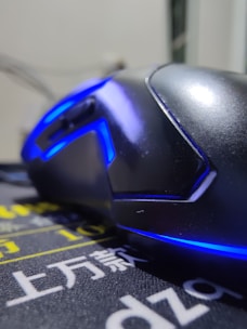 A close-up view of a sleek, black gaming mouse with blue LED lighting accents. The mouse is resting on a mouse pad with Asian characters and yellow and white text. The focus is on the curves and lighting of the mouse, giving it a futuristic and stylish appearance.