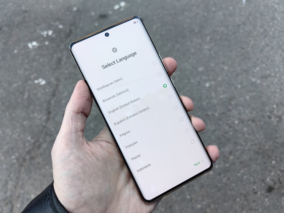 A hand holding a smartphone displaying a language selection screen with several language options including Azerbaijani, Bosnian, English, and others. The screen shows a light background with a circular selection indicator next to English.
