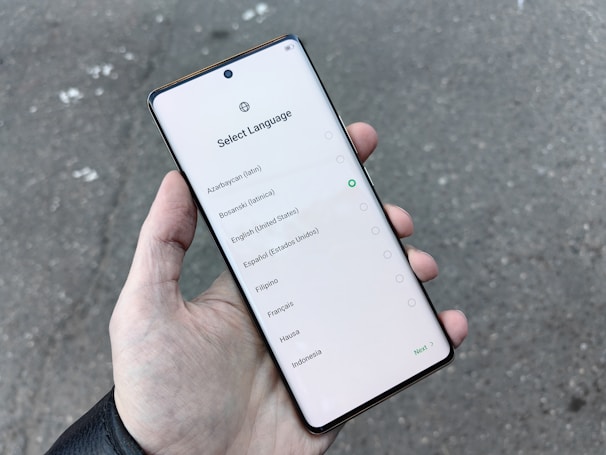 A hand holding a smartphone displaying a language selection screen with several language options including Azerbaijani, Bosnian, English, and others. The screen shows a light background with a circular selection indicator next to English.