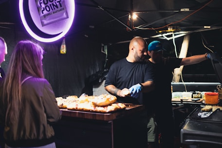 A food stall with two men preparing and grilling food under dim lighting. They are handling various food items, while a woman stands nearby, seemingly waiting. The scene is set in a dark environment with a sign above the stall and various cooking implements scattered around.
