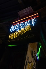 A neon sign with blue, red, and yellow lights is mounted on a dark background. The sign features text that appears to be stylized or artistic. Surrounding the sign, there is a grungy ambiance with some graffiti or weathered textures visible on the adjacent surfaces.
