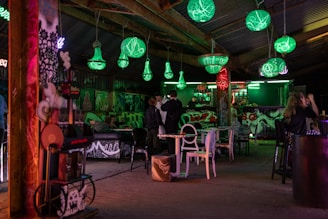 An indoor space decorated with green and red neon lights, featuring several spherical and chandelier-like light fixtures hanging from the ceiling. The walls are adorned with colorful graffiti art, and various mismatched chairs and tables are scattered throughout. People are gathered around the bar area, which is also artistically painted.