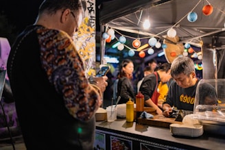 A bustling street food stall with a vendor preparing food. The stand is decorated with colorful paper lanterns. In the foreground, a person is capturing a photo on their smartphone. Various condiments are lined up on the counter.