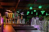 A dimly lit bar scene with neon signs displaying words like 'wonderful' and 'delicious' above a stocked bar. Patrons stand and sit around, socializing. Green chandeliers hang from the ceiling, casting light on the surroundings. The atmosphere is colorful and vibrant with a mix of industrial and artistic elements.