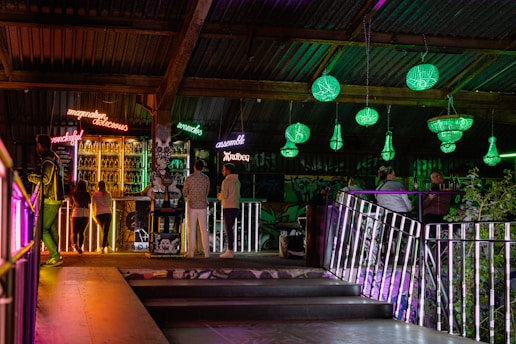 A dimly lit bar scene with neon signs displaying words like 'wonderful' and 'delicious' above a stocked bar. Patrons stand and sit around, socializing. Green chandeliers hang from the ceiling, casting light on the surroundings. The atmosphere is colorful and vibrant with a mix of industrial and artistic elements.
