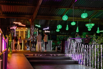 A dimly lit bar scene with neon signs displaying words like 'wonderful' and 'delicious' above a stocked bar. Patrons stand and sit around, socializing. Green chandeliers hang from the ceiling, casting light on the surroundings. The atmosphere is colorful and vibrant with a mix of industrial and artistic elements.