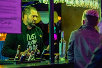 Two people are engaged in conversation at a bar. One person, wearing a dark jacket with a distinctive logo, is speaking while the other, with a back view, listens attentively. Several bottles are on the counter, illuminated by multicolored neon lights and surrounded by hanging glasses.