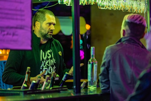 Two people are engaged in conversation at a bar. One person, wearing a dark jacket with a distinctive logo, is speaking while the other, with a back view, listens attentively. Several bottles are on the counter, illuminated by multicolored neon lights and surrounded by hanging glasses.