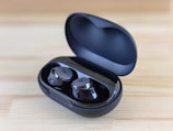 A pair of sleek black wireless earbuds resting in an open charging case on a wooden surface. The earbuds have a shiny finish with a logo on each, and the case is slightly tilted to display the earbuds and the inner compartment.