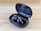 A pair of sleek black wireless earbuds resting in an open charging case on a wooden surface. The earbuds have a shiny finish with a logo on each, and the case is slightly tilted to display the earbuds and the inner compartment.