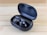 A pair of sleek black wireless earbuds resting in an open charging case on a wooden surface. The earbuds have a shiny finish with a logo on each, and the case is slightly tilted to display the earbuds and the inner compartment.