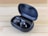 A pair of sleek black wireless earbuds resting in an open charging case on a wooden surface. The earbuds have a shiny finish with a logo on each, and the case is slightly tilted to display the earbuds and the inner compartment.