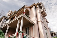 A large, elegant house is adorned with festive red and green decorations, including wreaths and ribbons. The house features detailed architectural elements such as columns and railings, and is set against a partly cloudy sky. The building has a pastel pink facade with multiple balconies.