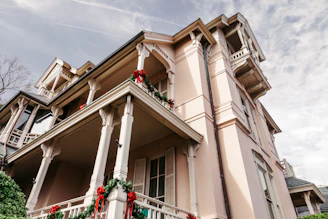 A large, elegant house is adorned with festive red and green decorations, including wreaths and ribbons. The house features detailed architectural elements such as columns and railings, and is set against a partly cloudy sky. The building has a pastel pink facade with multiple balconies.