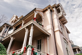 A large, elegant house is adorned with festive red and green decorations, including wreaths and ribbons. The house features detailed architectural elements such as columns and railings, and is set against a partly cloudy sky. The building has a pastel pink facade with multiple balconies.
