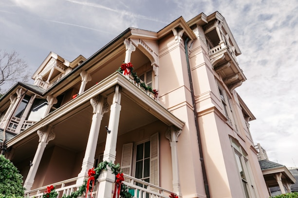 A large, elegant house is adorned with festive red and green decorations, including wreaths and ribbons. The house features detailed architectural elements such as columns and railings, and is set against a partly cloudy sky. The building has a pastel pink facade with multiple balconies.