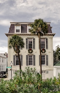A historic three-story building with a slate roof and decorative trim has large windows with shutters. Two palm trees stand in front, flanking the entrance. Wreaths are hung above the windows, suggesting a festive atmosphere. A street sign and vintage-style lamp are visible on the corner, next to some bushes.