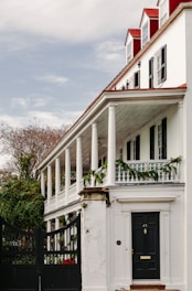 A large, white colonial-style house with a red roof and multiple chimneys is shown. The house features a prominent front porch supported by white columns and decorated with green garlands. There are several tall windows with black shutters and a black door with an address number on it. Surrounding the house, there is greenery and a black iron gate at the entrance.
